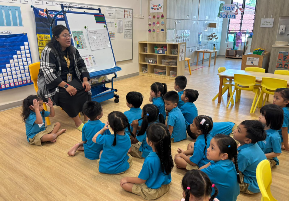 Teacher sits by a whiteboard speaking to a group of young children in blue uniforms seated on the floor.